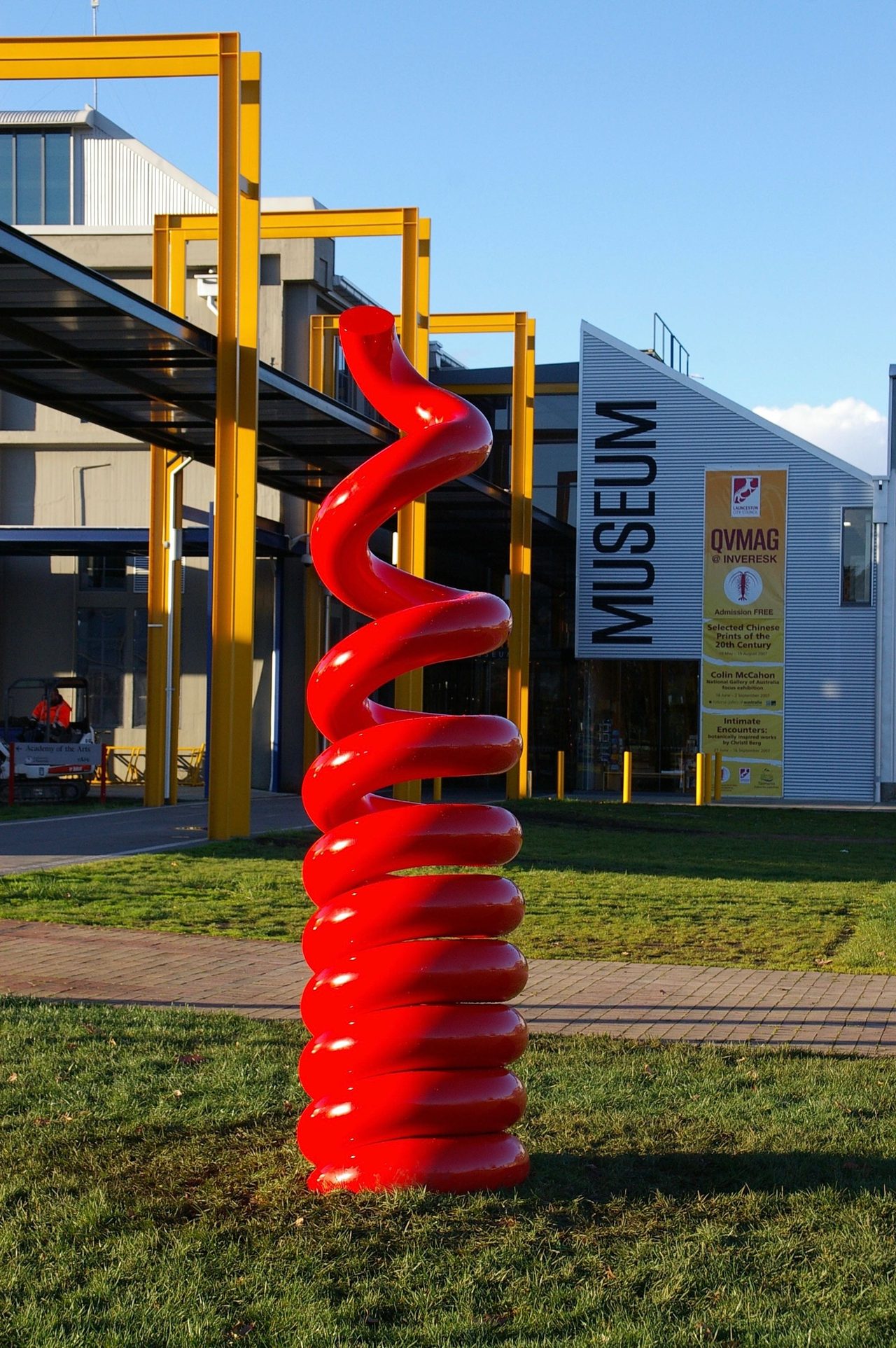 Earth Drill, a red painted steel spiral sculpture rising from a lawn in front of the QVMAG museum entrance sign, by David Hamilton
