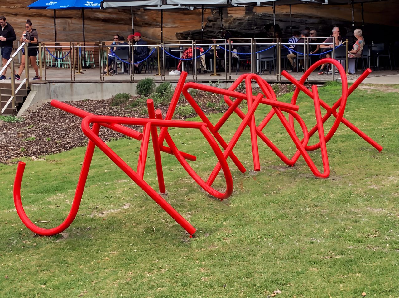 Jackinabox, a large tangled red painted steel line sculpture standing on a lawn beside an outdoor cafe, by David Hamilton