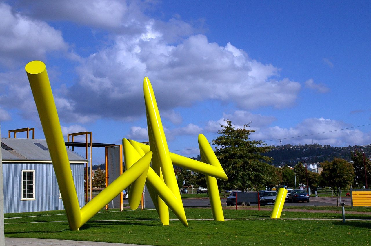 Bolt, a cluster of angular yellow steel rods on grass beside a road, with hills in the background, by David Hamilton