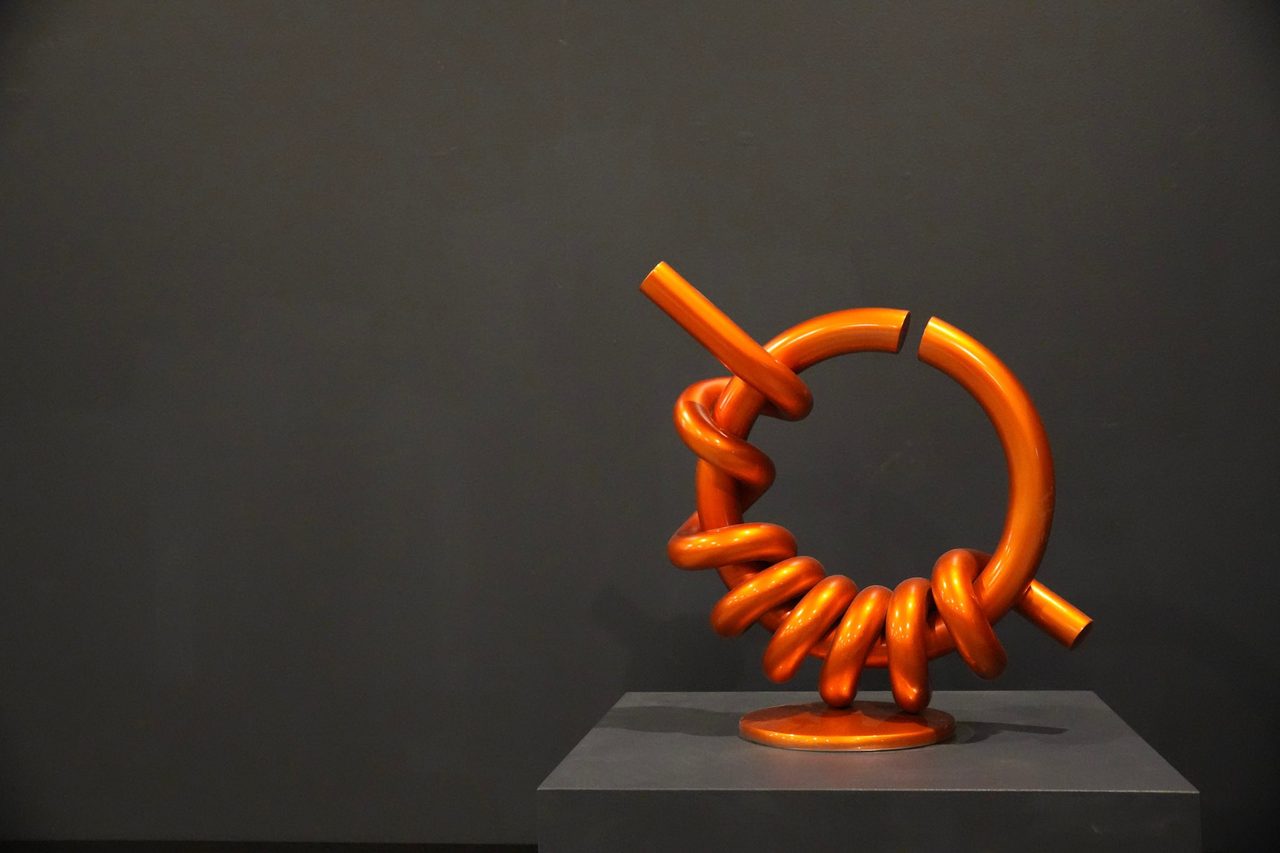 Hot Sun, an orange painted steel ring wrapped in coiled steel rod, on a low plinth against a dark wall, by David Hamilton
