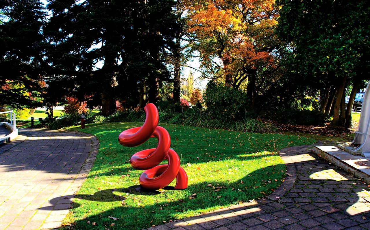 A red painted steel spiral sculpture, tapering to a point at its top, standing in an autumn garden, by David Hamilton