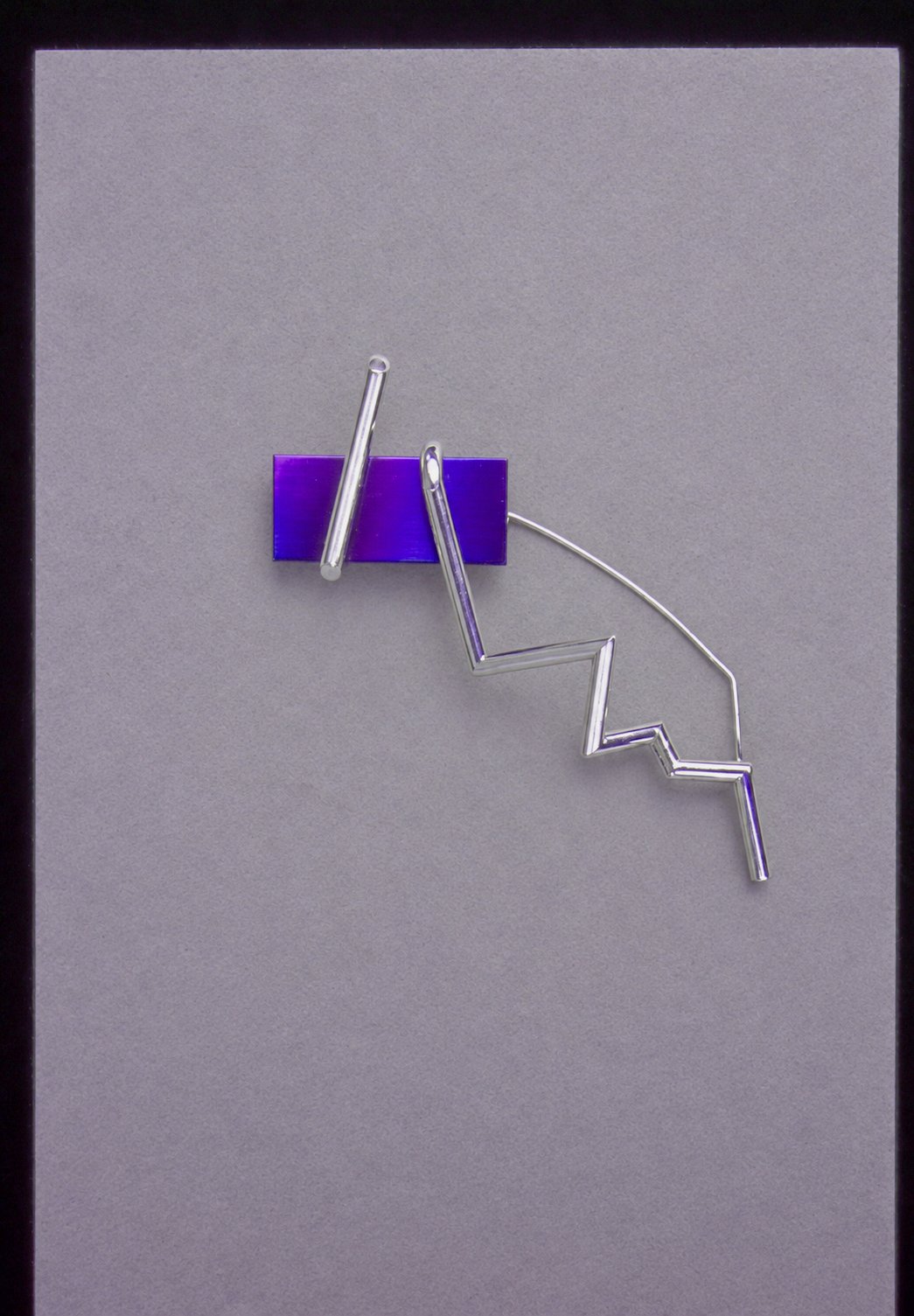 A silver brooch of a zigzag lightning-bolt wire pinned across a dark blue rectangular bar, by David Hamilton