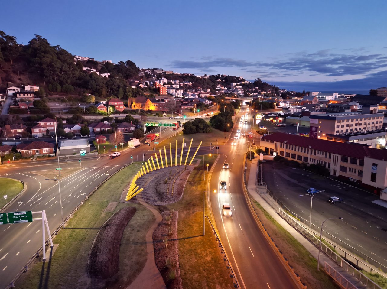 Welcome Gesture at dusk from above: a line of leaning yellow poles beside a highway into Burnie, with the town lit behind