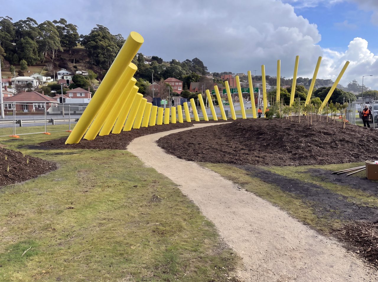 Welcome Gesture, a row of tall leaning yellow steel poles along a curved gravel path in Burnie, by David Hamilton
