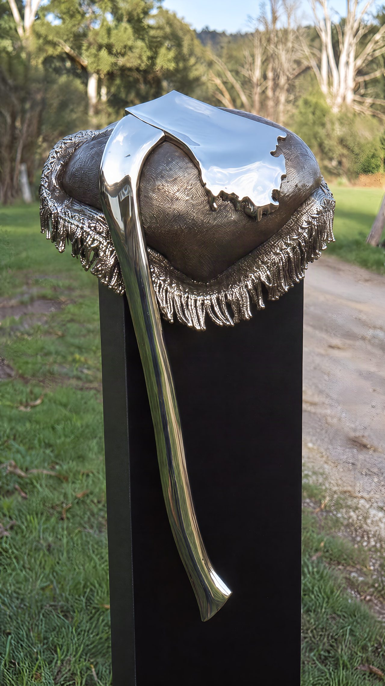 A polished silver axe blade set in a fringed cushion-like mount, mounted on a black outdoor plinth among trees, by David Hamilton