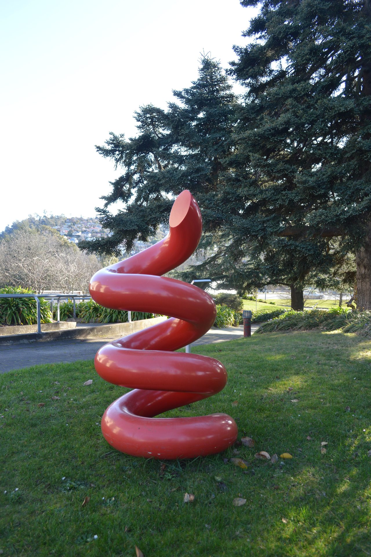 Earth Drill, a tall red painted steel spiral standing on grass in front of pine trees at the Queen Victoria Museum and Art Gallery, Launceston, by David Hamilton